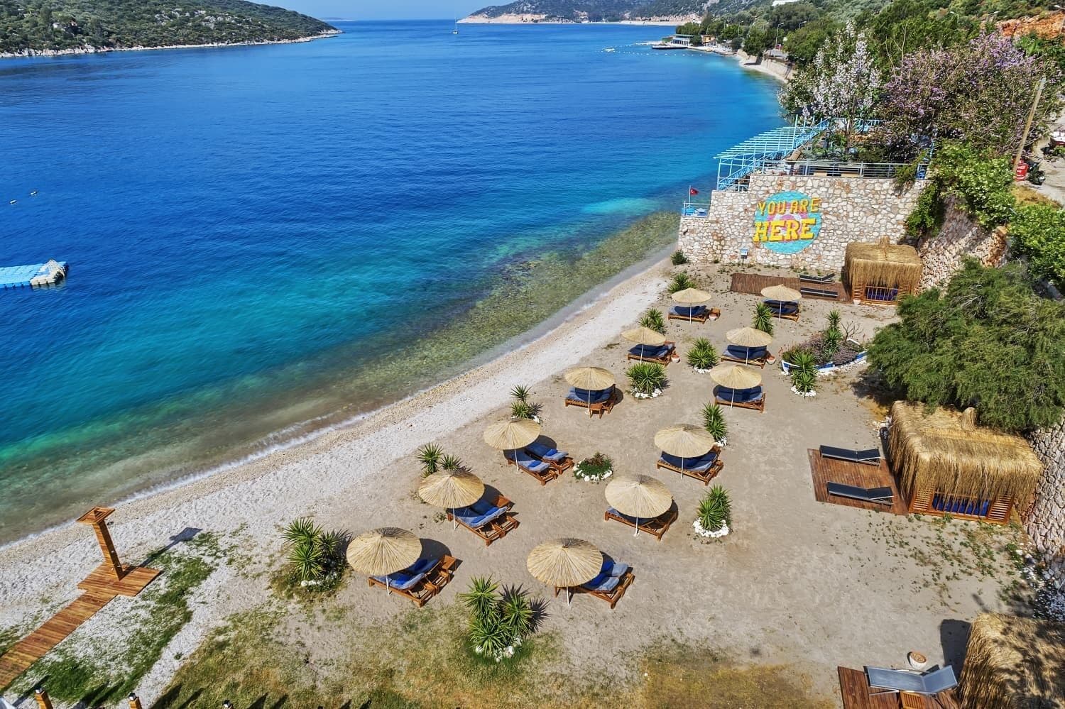 Ein Hotel am Strand mit Liegestühlen und Sonnenschirmen, eine Steinmauer mit einem Schild und Blick auf das Meer und die Hügel.