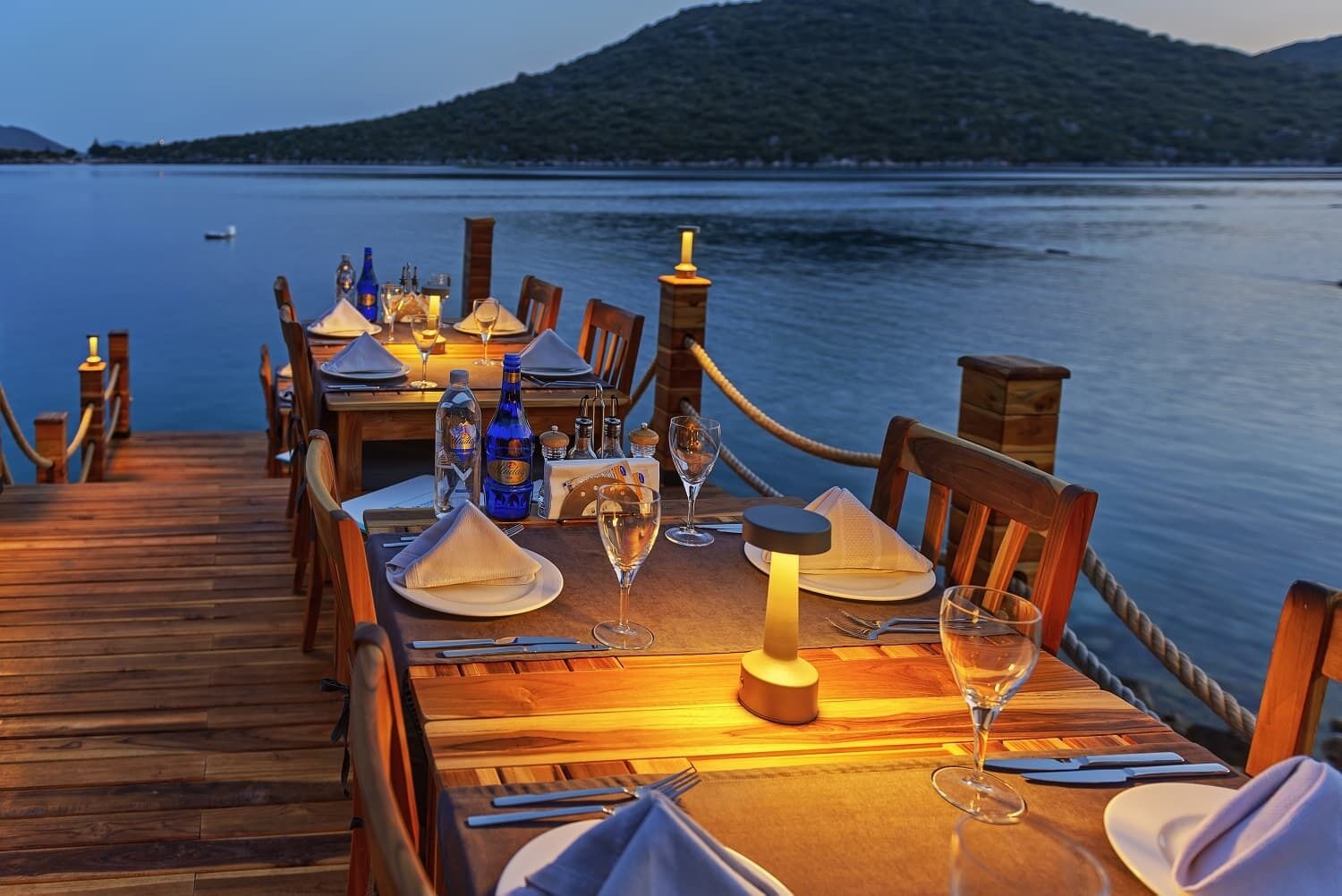 Una terraza de madera con una mesa preparada para comer, con vistas a un cuerpo de agua tranquilo y un paisaje montañoso.