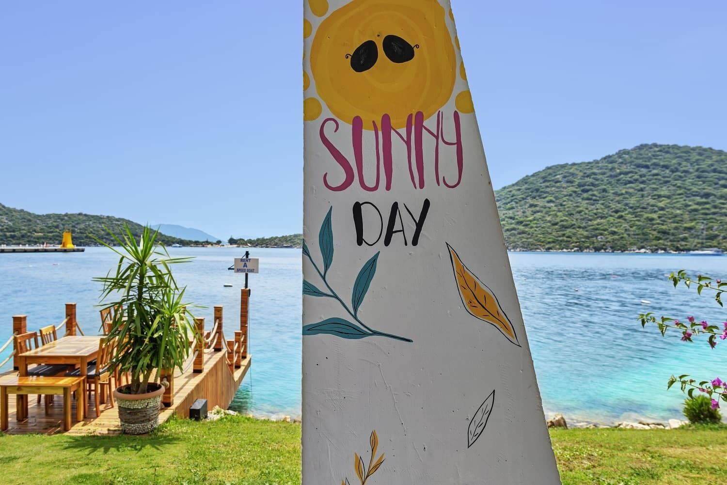 A wooden pier with a table and chairs, a sign reading "SUNNY DAY," and a view of a calm lake with a mountain in the background.