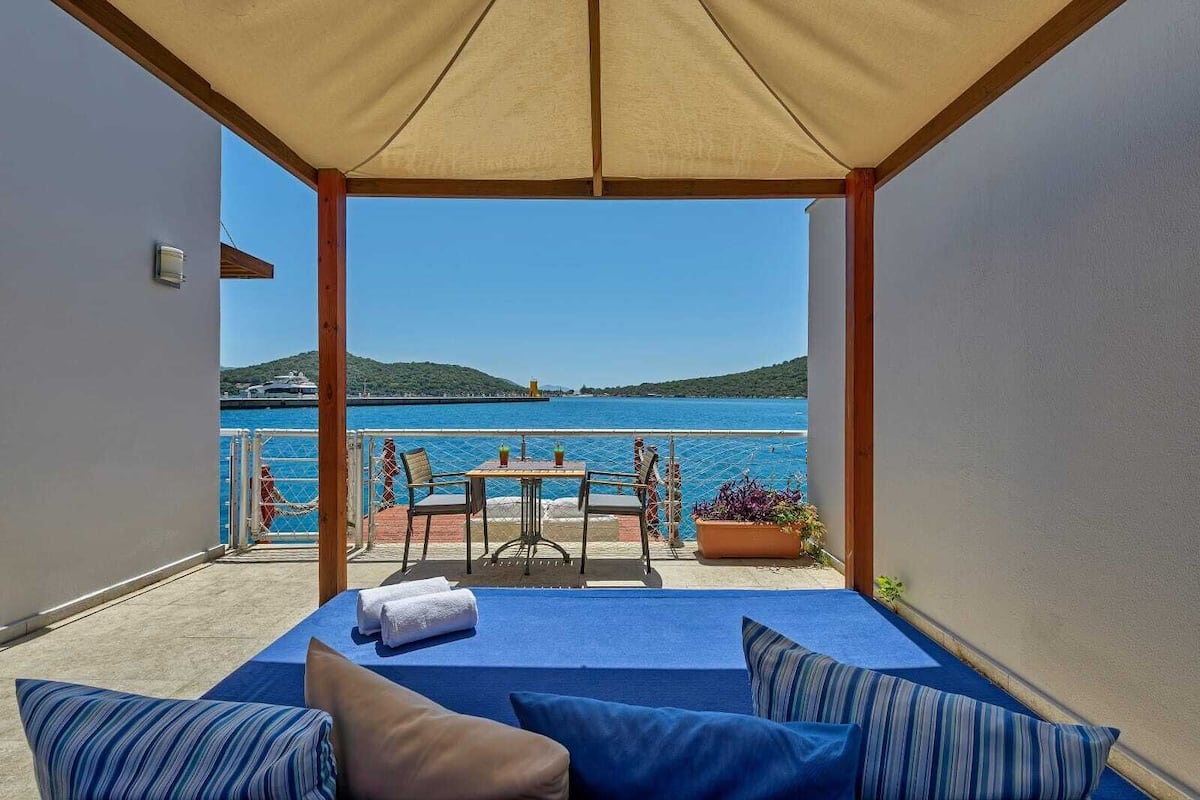 A balcony with a view of the sea, a table with chairs, and a sofa with cushions.