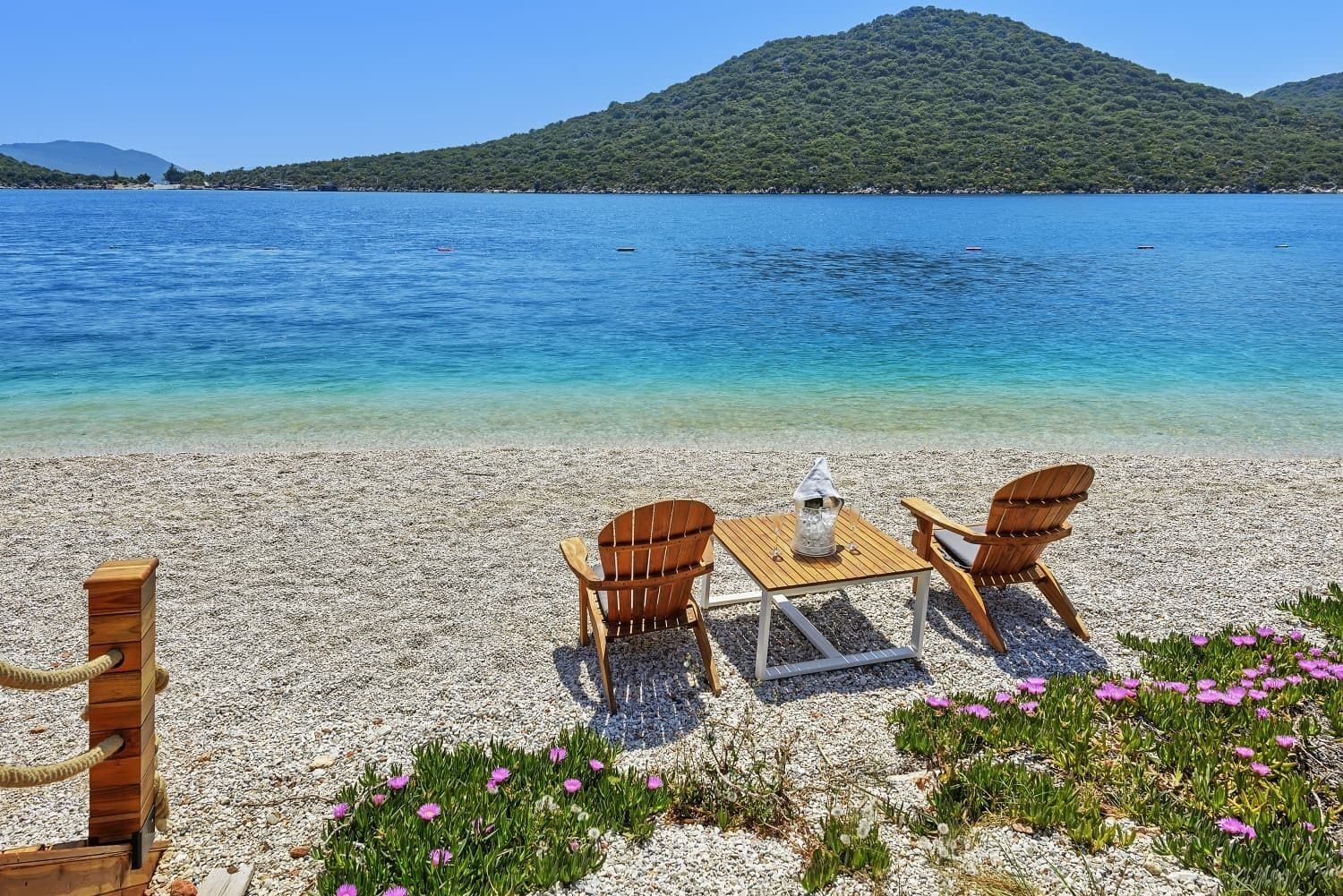A beach with a wooden table and chairs, a small wooden structure, and a clear view of the sea and a hill in the background.