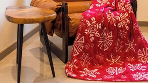 Uma poltrona de couro com uma mesinha de madeira, um cobertor estampado em vermelho e uma janela com vista para um edifício.
