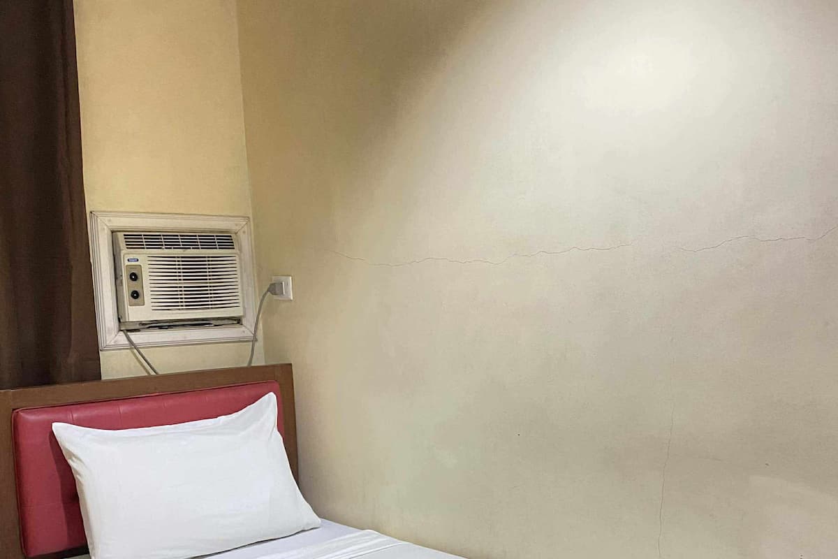 Una cama individual con ropa blanca, cabecera roja y almohada blanca. Una unidad de aire acondicionado está montada en la pared sobre la cama.