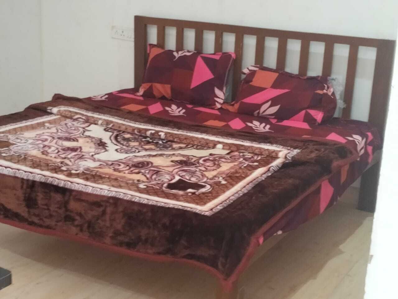 A bed with a patterned bedspread and pillows, a wooden headboard, and a visible electrical outlet on the wall.