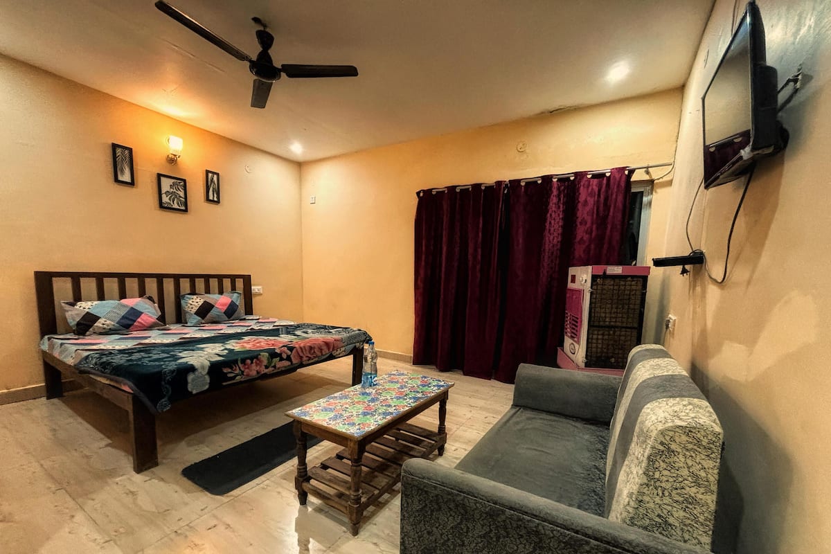 Un dormitorio con cama, sofá, una mesita, ventilador de techo, televisor montado en la pared y ventana con cortinas.