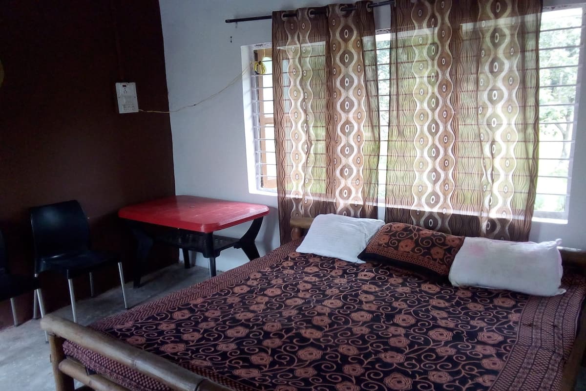 Una habitación con una cama, una mesa roja, dos sillas y una ventana con cortinas estampadas.