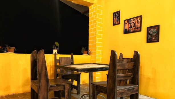 Un área de comedor al aire libre con mobiliario de madera y un pilar de ladrillo, frente a una pared amarilla con obras de arte.