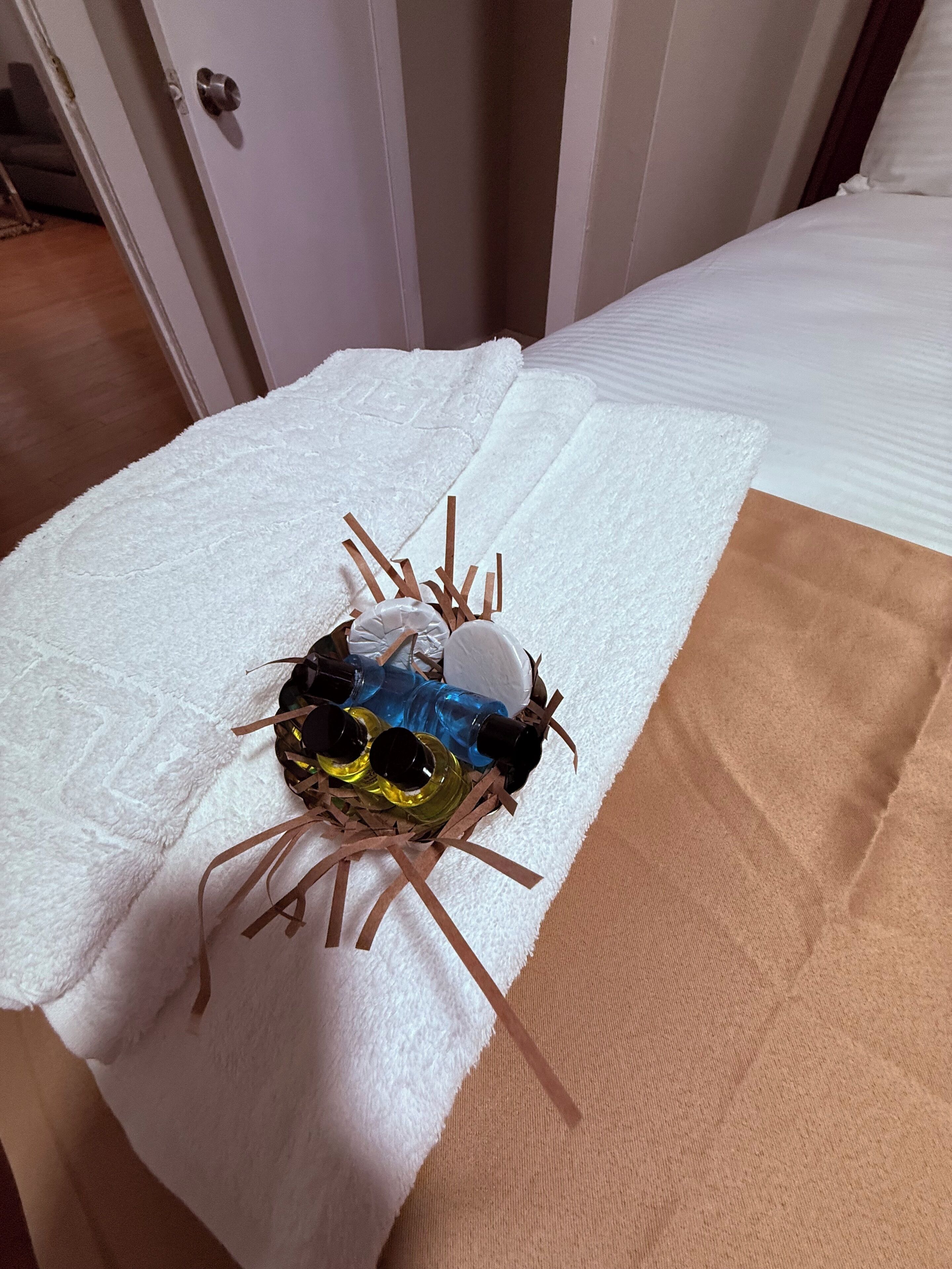 Un lit avec une couverture blanche et un drap marron, une serviette blanche, et un panier contenant des bougies et des bouteilles, posé sur le lit.