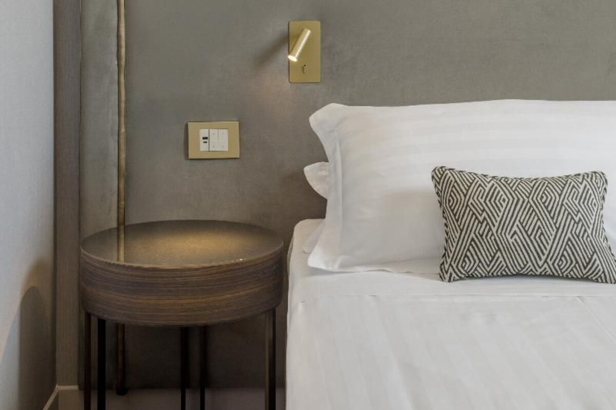 A neatly made bed with white linens, a patterned pillow, a small round table, and a wall-mounted lamp.