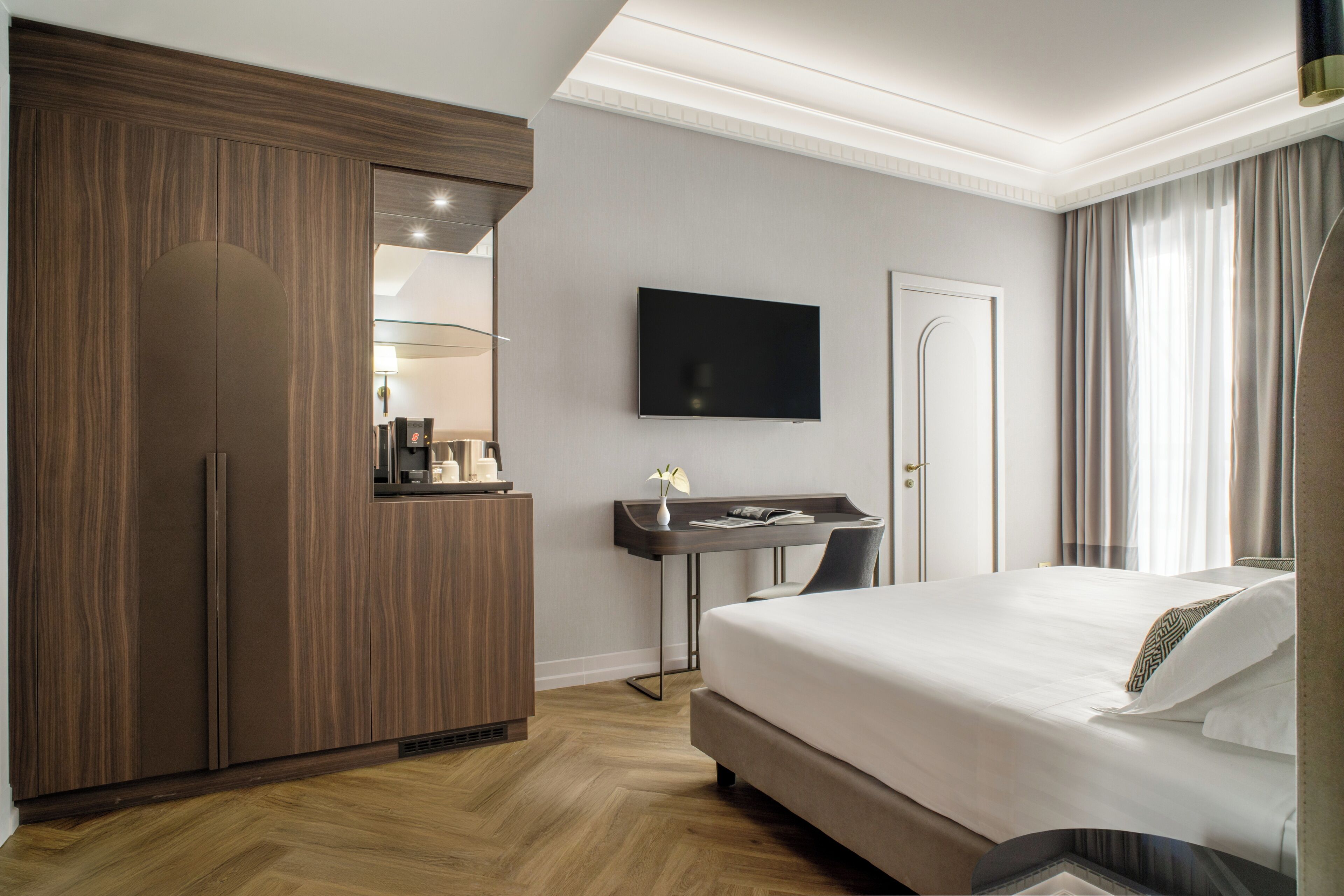 A hotel room with a large bed, a desk with a chair, a TV mounted on the wall, and a wardrobe.