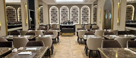 Un restaurante con varias mesas y sillas, una barra central y amplias ventanas en arco que muestran estantes con objetos.