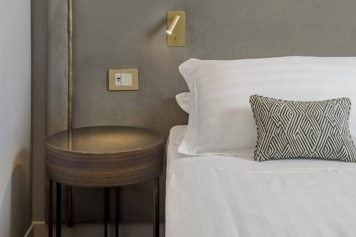 Un lit bien fait, avec du linge de lit blanc, un coussin à motifs, une petite table ronde et une lampe fixée au mur.