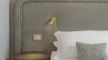 Un letto rifatto con lenzuola bianche e un cuscino decorato, uno sgabello rotondo in legno, una lampada a parete e un interruttore sulla parete.