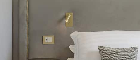 A neatly made bed with white bedding and a patterned pillow, a round wooden stool, a wall-mounted lamp, and a light switch on the wall.