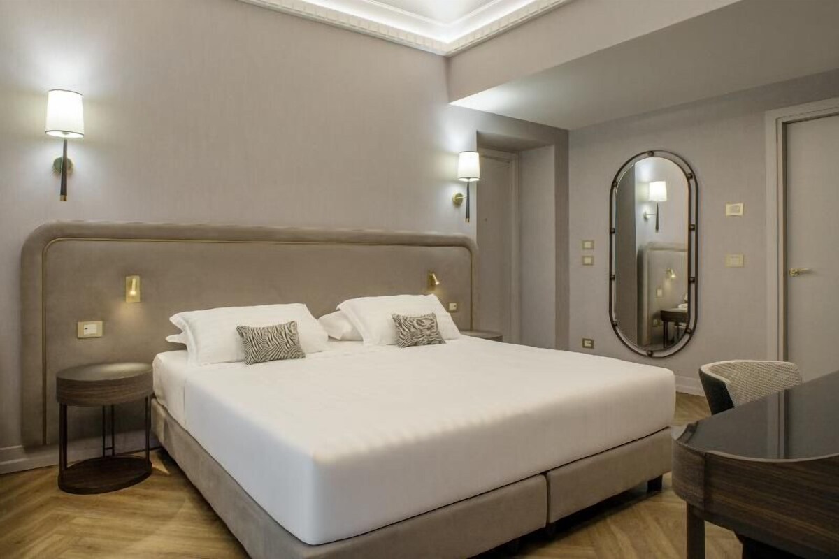 Una habitación de hotel con una cama grande, mesitas de noche, un escritorio y un espejo.
