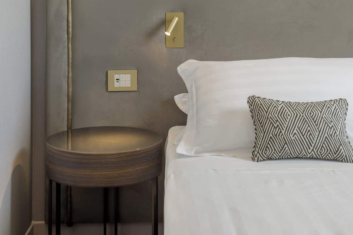 Un lit bien fait, avec une literie blanche et un coussin à motifs, un tabouret rond en bois, une lampe fixée au mur et un interrupteur sur le mur.