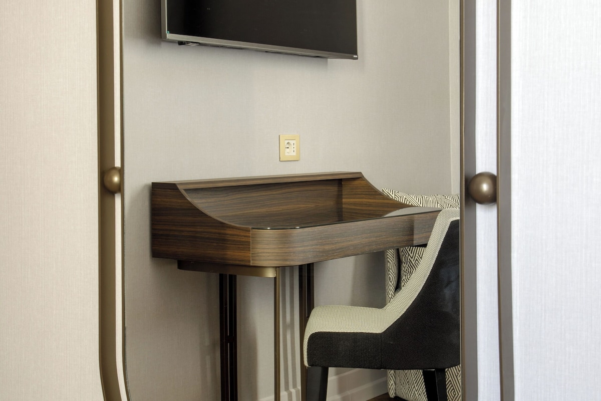 Une pièce avec un miroir rond, un bureau, une chaise et un téléviseur fixé au mur.