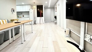 A modern kitchen with white cabinets, a wooden dining table, and a large flat-screen TV mounted on the wall. - 5 mins to Unseo Station, the nearest to the Incheon Airport, Located in Cafe St. (Incheon)
