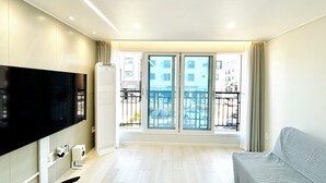 A modern living room with a flat-screen TV, a standing heater, a sofa, and a balcony with a view of buildings. - 5 mins to Unseo Station, the nearest to the Incheon Airport, Located in Cafe St. (Incheon)