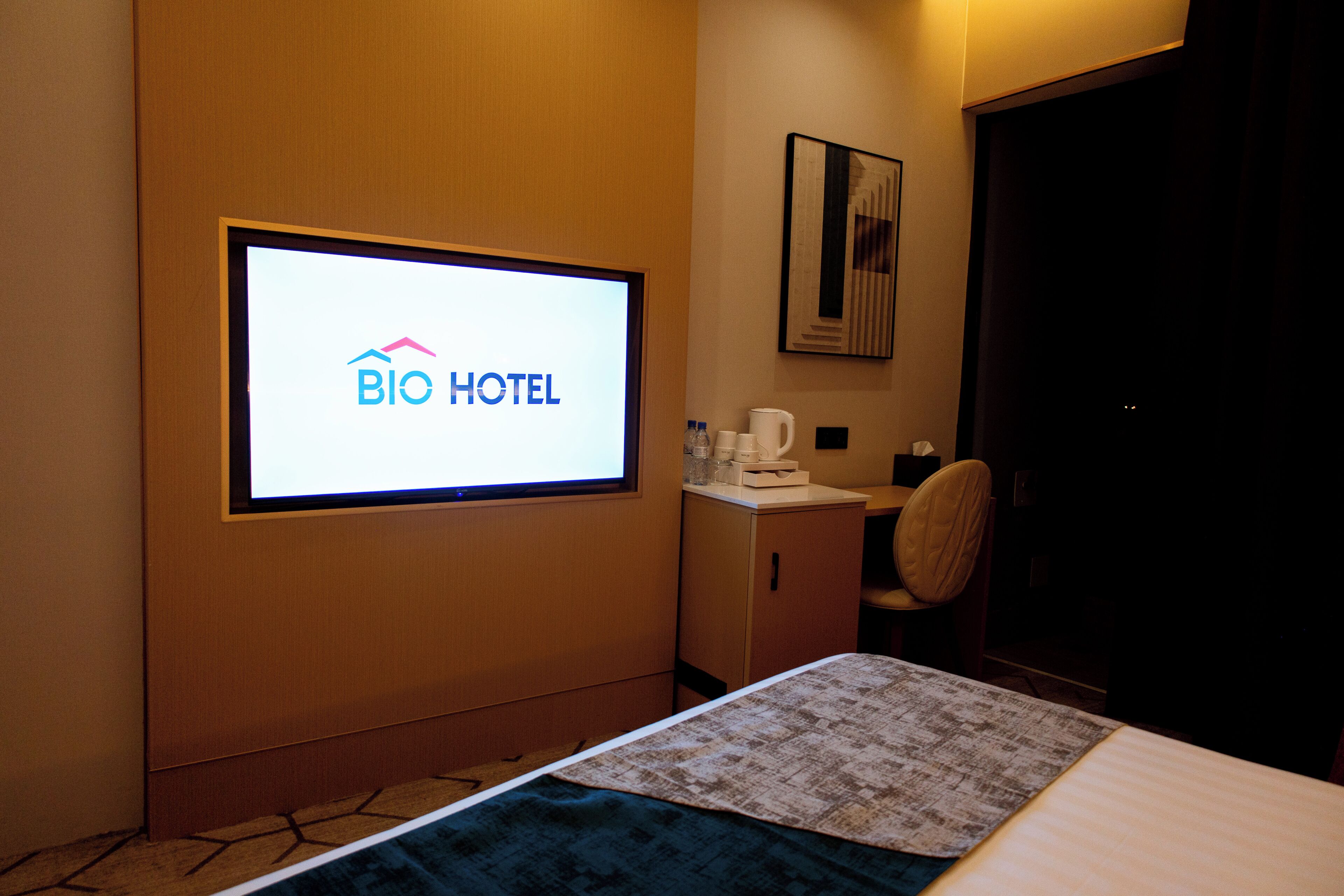 A hotel room with a bed, a desk, a chair, a coffee maker, and a screen displaying the hotel's logo.
