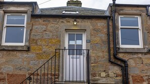 A stone building with a white door, a small balcony with a black railing, and two windows on either side of the door. - Rocklands (No 11) | Elie (Elie)