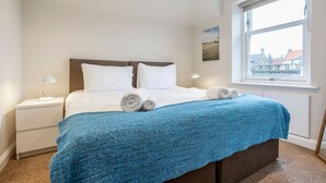 A neatly made bed with white linens and a blue blanket, a bedside lamp, and a window with a view of buildings. - Rocklands (No 11) | Elie (Elie)