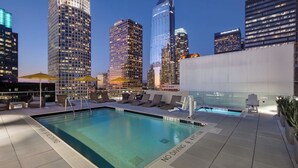 Una piscina en la azotea con sillones y sombrillas, rodeada de edificios altos.