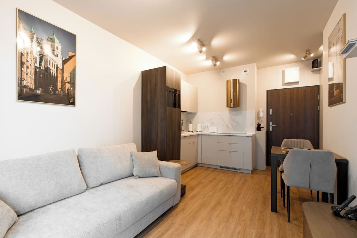 Un apartamento moderno con un sofá gris, suelo de madera, una cocina con armarios blancos y un comedor con una silla.