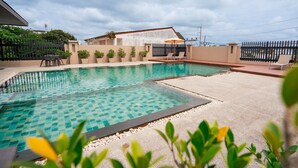 A swimming pool with a checkered pattern, surrounded by a low fence, and a building with a sloped roof in the background. - Pink Residence Chalong (Rawai)