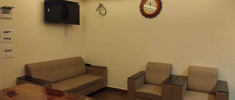 Uma sala de estar moderna com um sofá, duas poltronas, uma mesa de centro de madeira, uma TV de tela plana na parede, um ventilador de teto e um relógio.