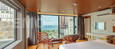A hotel room with a large bed, two wicker chairs, a small table, a TV, and a view of the ocean and buildings.