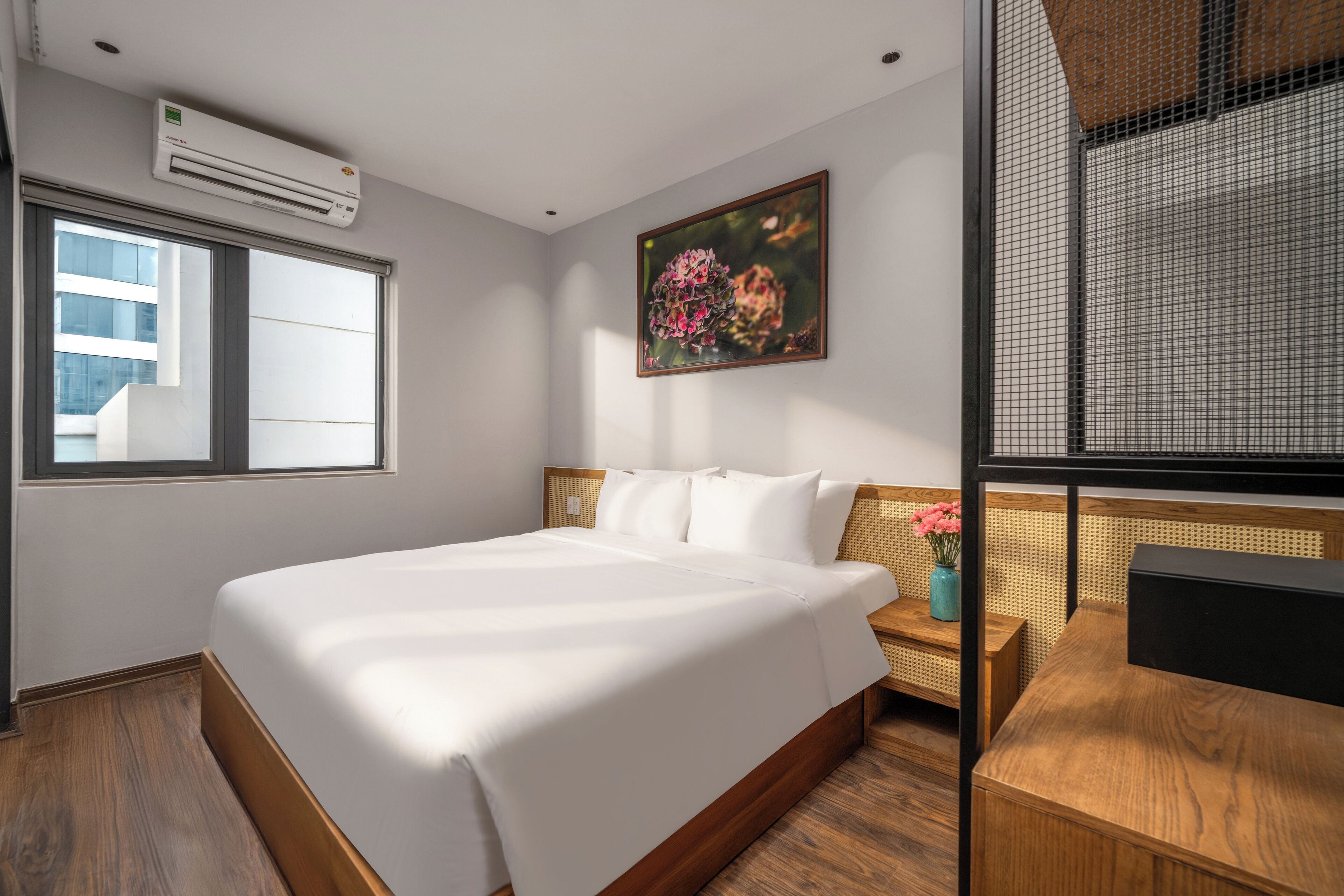 A hotel room with a large bed, a window, a painting, a desk, and an air conditioning unit.