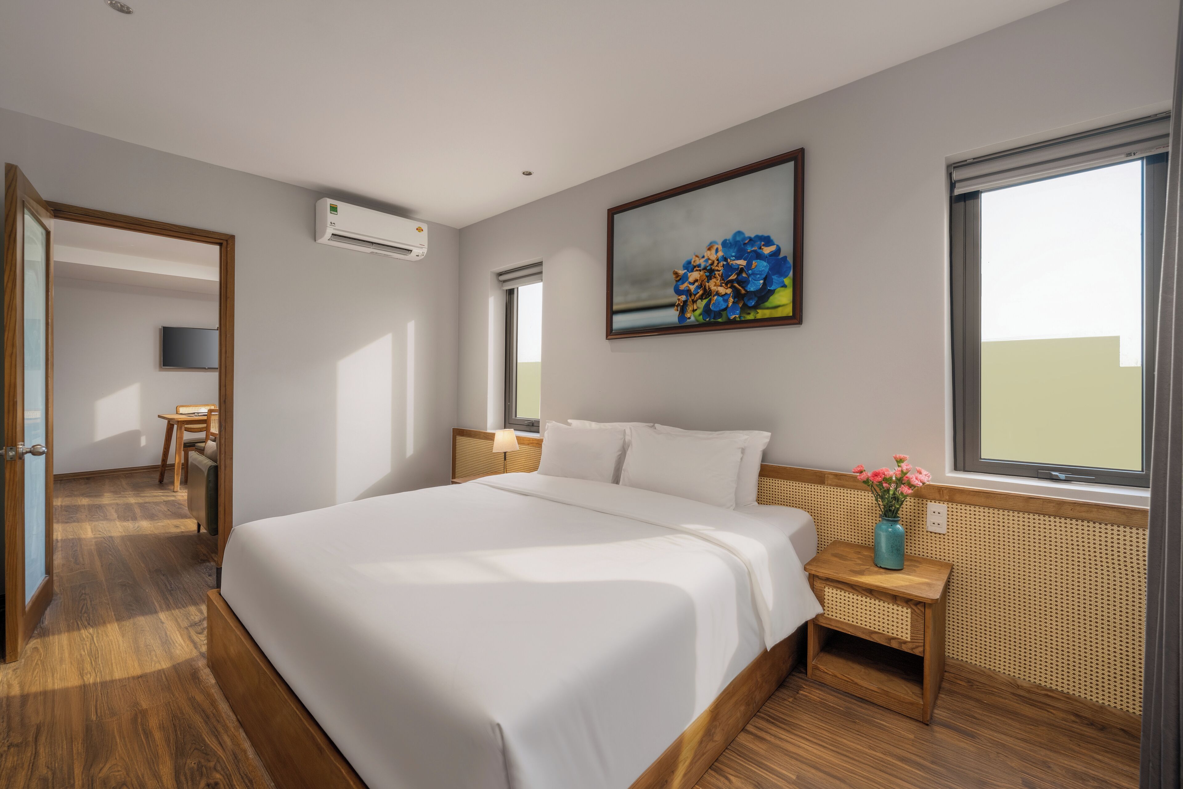 Une chambre d’hôtel moderne avec un grand lit, une petite table de chevet ornée d’un vase de fleurs, un climatiseur fixé au mur et un tableau au-dessus du lit.