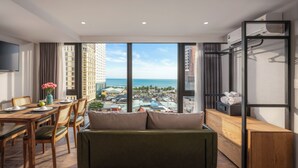 Quarto de hotel moderno com uma janela grande que oferece vista para a cidade, um espaço de refeições com mesa e cadeiras de madeira, e uma área de estar com sofá e uma televisão pequena em um suporte.