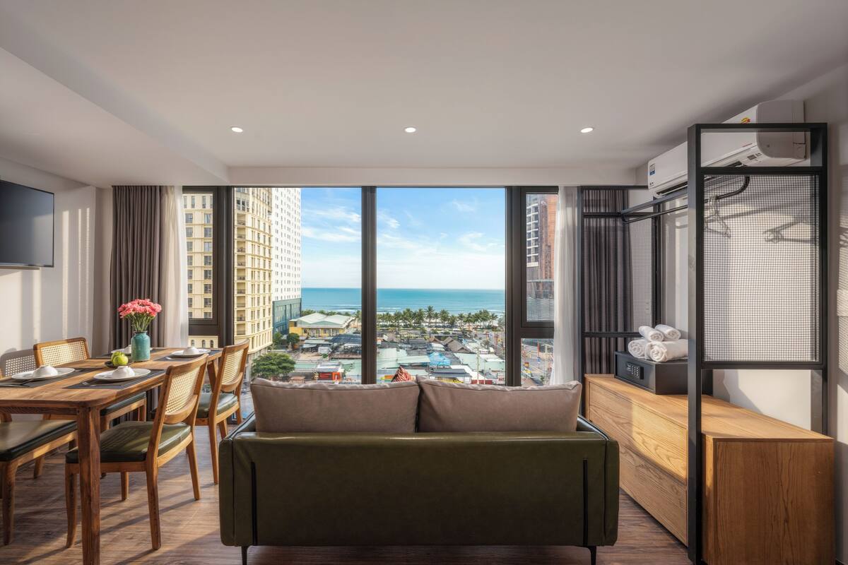 Een moderne hotelkamer met een groot raam dat uitzicht geeft op de stad, een eethoek met een houten tafel en stoelen, en een zithoek met een bank en een kleine televisie op een standaard.