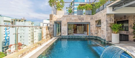 An indoor swimming pool with a waterfall feature, surrounded by modern buildings and balconies with plants.