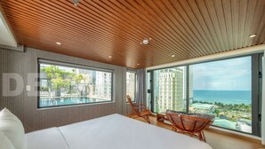 Quarto de hotel moderno com uma cama grande, área de jantar com cadeiras de vime e varanda com vista para o oceano e edifícios.