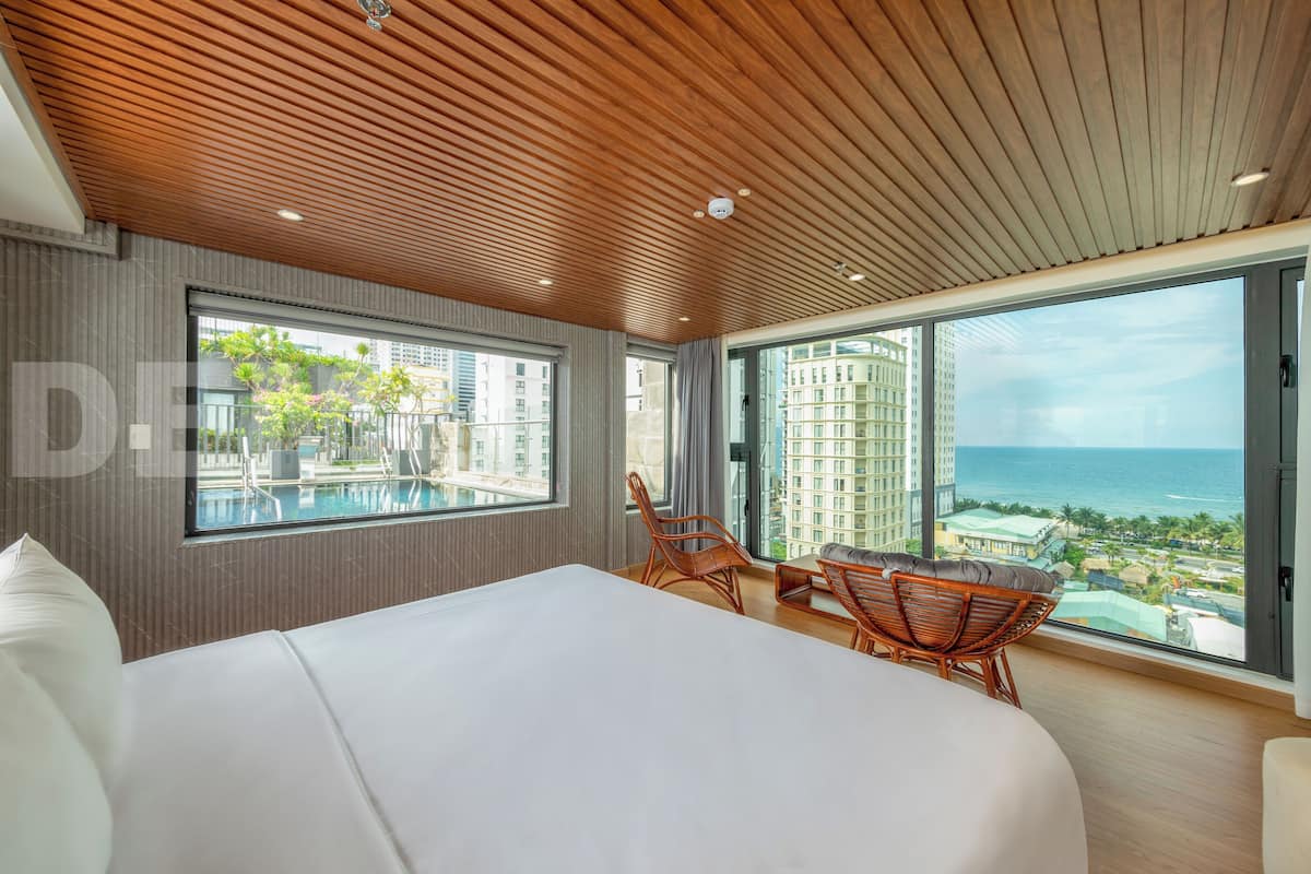 Een moderne hotelkamer met een groot bed, een zithoek met rieten stoelen, en een balkon met uitzicht op de zee en gebouwen.