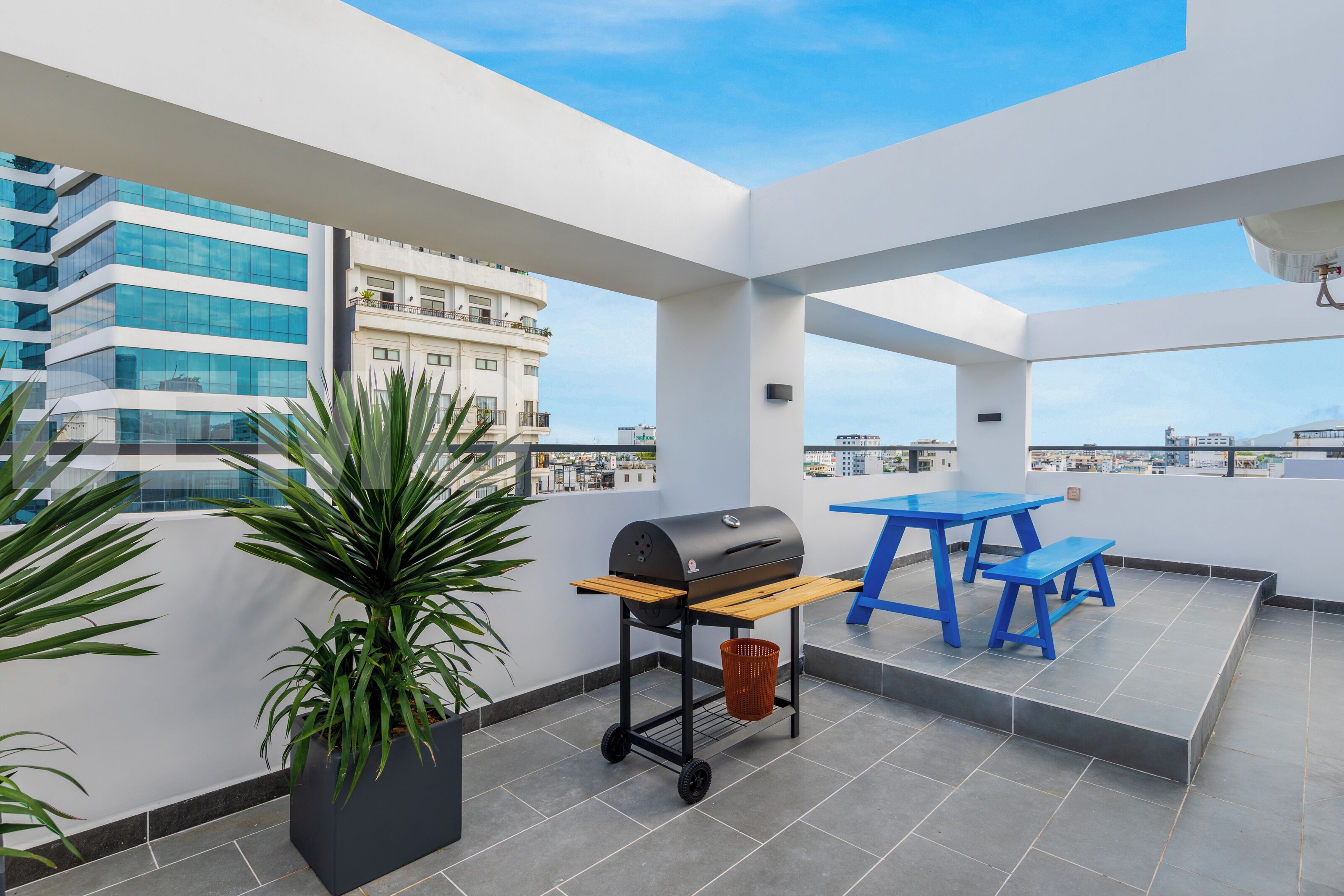 A rooftop terrace with a grill, blue table, and potted plants.