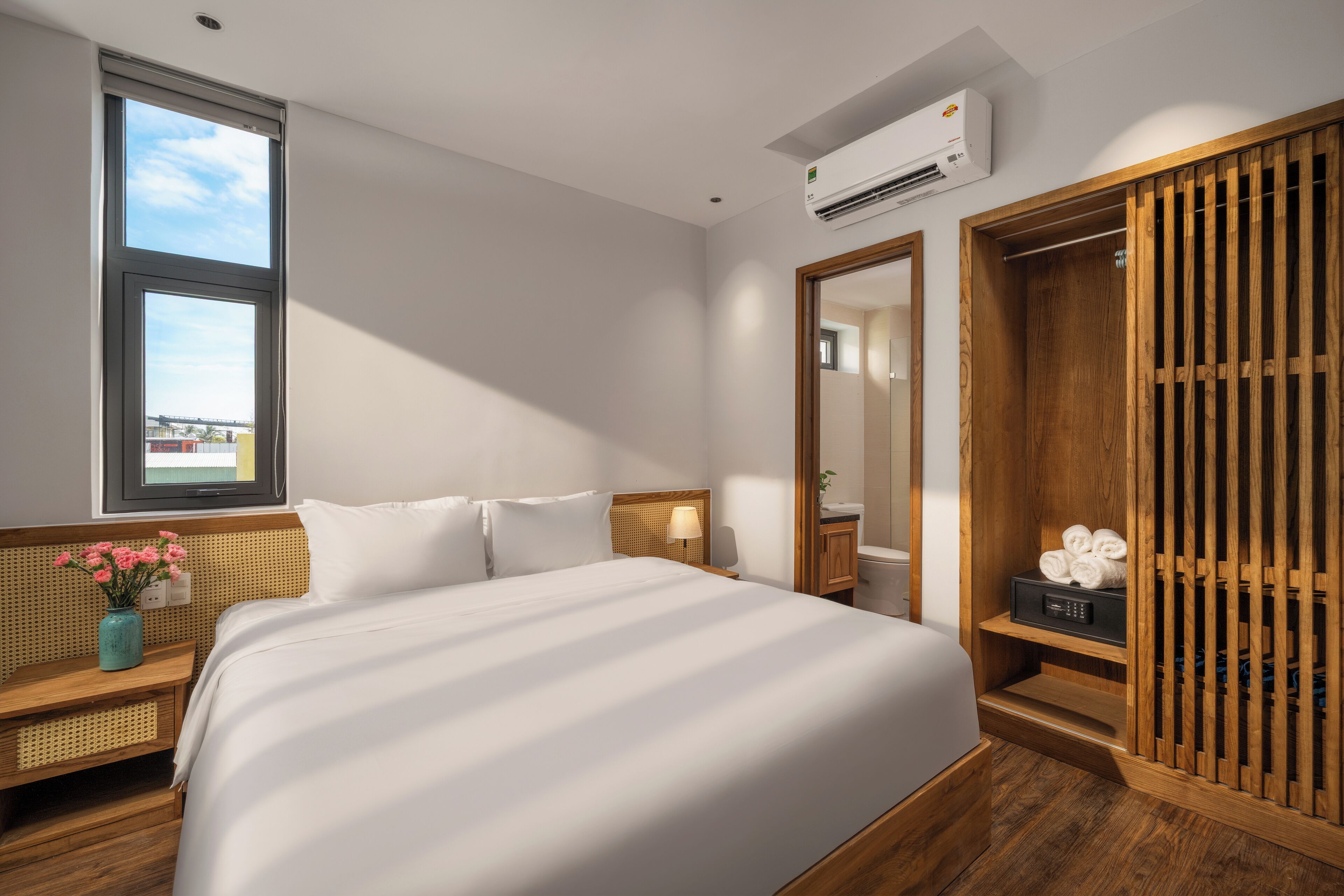 Ein modernes Hotelzimmer mit einem großen Bett, einem Nachttisch mit Lampe, einem Holzschrank und einem Fenster mit Blick.