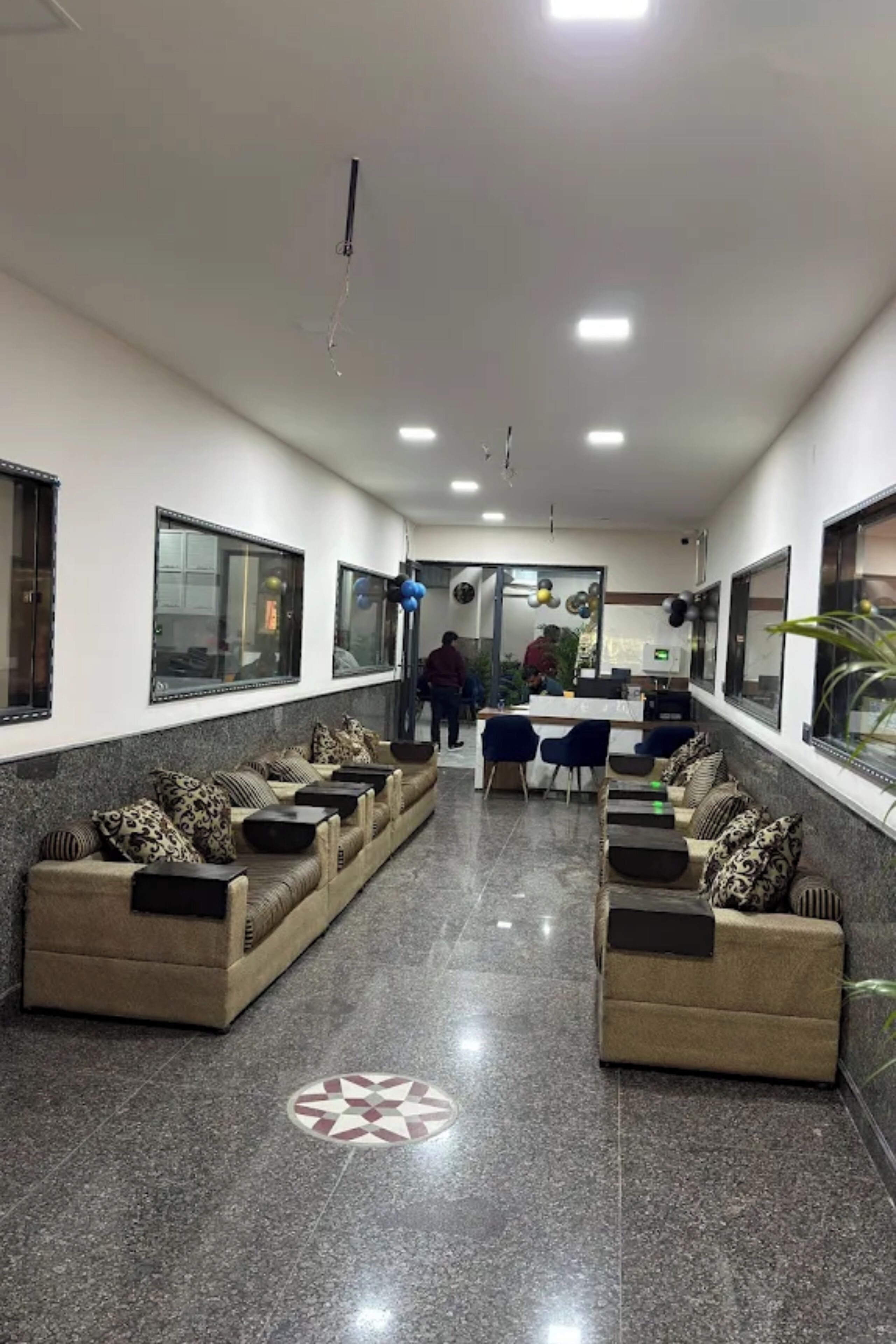 A reception area with a long, narrow layout, featuring a reception desk, two sofas with patterned cushions, and a tiled floor with a geometric pattern.