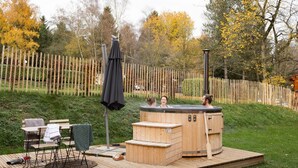 Een hottub op een houten terras met treden, omringd door een houten hek en bomen met herfstbladeren.