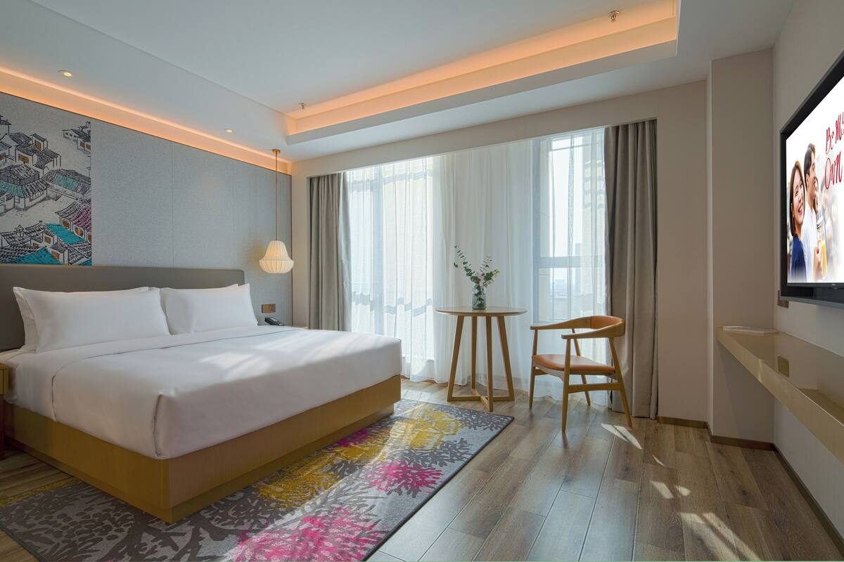 A hotel room with a large bed, a small table with a chair, a TV, and a large window with curtains.