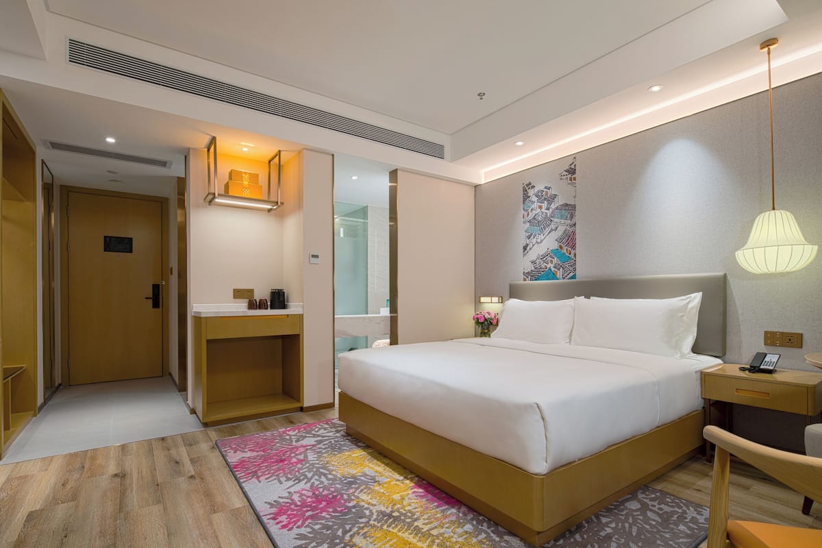 A hotel room with a large bed, a bedside table with a lamp, a small desk, a mirror, and a shower area.
