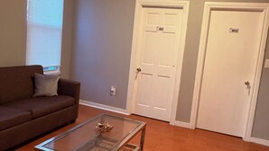 Un salón con un sofá marrón, una mesa de centro de vidrio, dos puertas blancas y una lámpara en el techo.