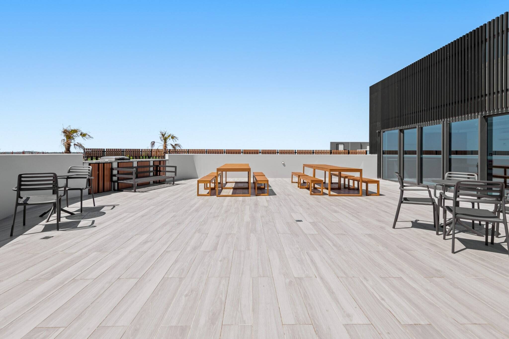 Una terraza en la azotea con mesas y sillas de madera, una valla de madera y un edificio moderno con grandes ventanales.