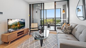 Un salon moderne doté d’un téléviseur à écran plat, d’une table basse en verre, d’un canapé gris et offrant une vue sur un balcon aménagé avec du mobilier d’extérieur.