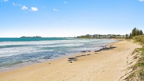 A beach with clear skies, calm waters, and a distant shoreline with buildings. - Beachside 2-Bed with Pools, Gym, BBQ and Parking (Alexandra Headland)