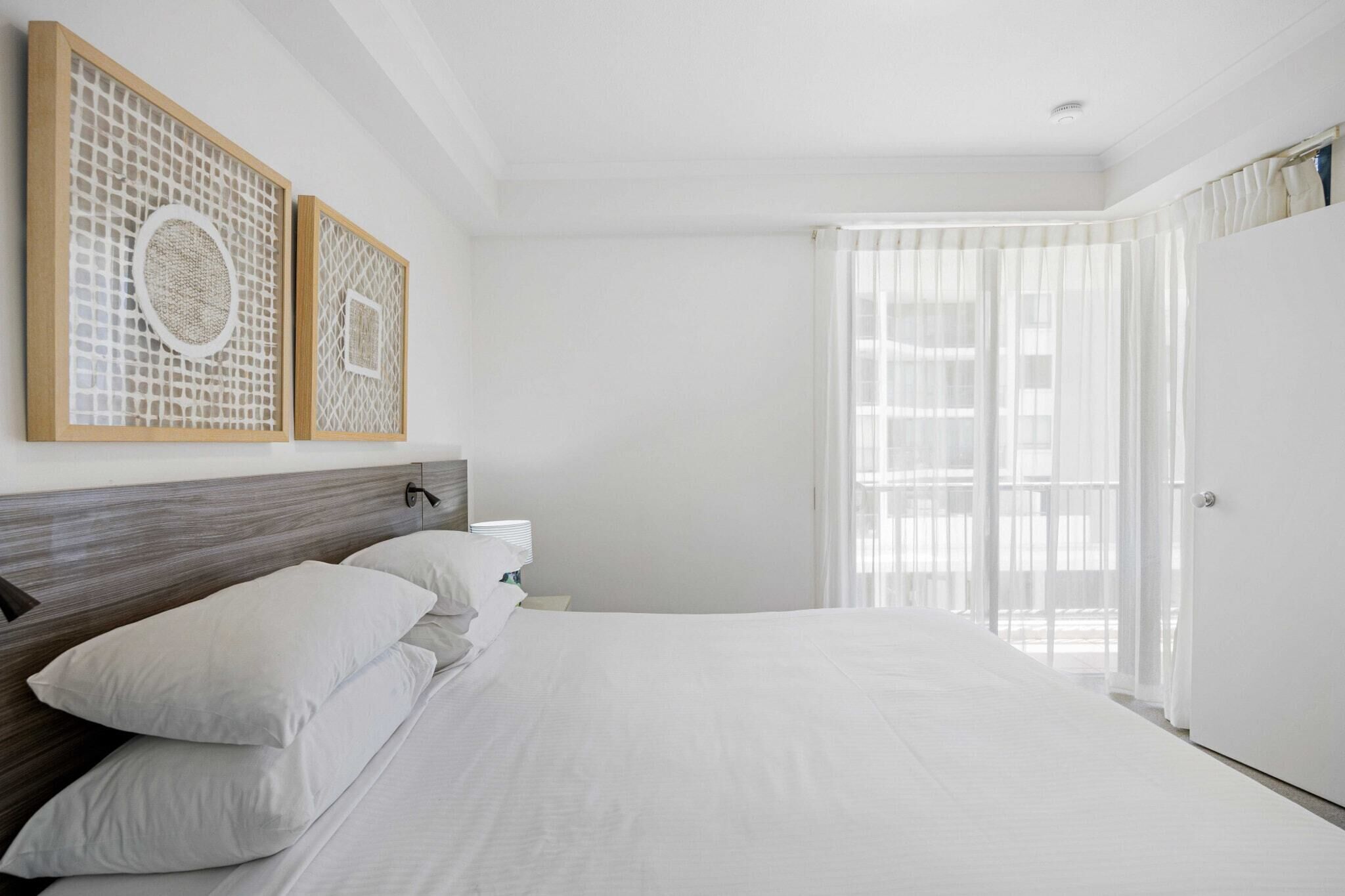 A hotel room with a large bed, a wooden headboard, two wall-mounted decorative panels, a bedside table with a lamp, and a large window with sheer curtains.
