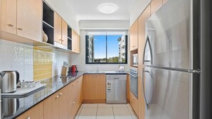 A modern kitchen with wooden cabinets, a stainless steel refrigerator, and a window with a view of buildings. - Beachside 2-Bed with Pools, Gym, BBQ and Parking (Alexandra Headland)
